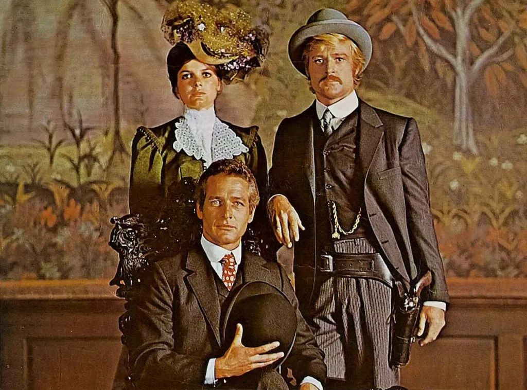 Three individuals in sophisticated attire from the late 19th or early 20th century. A woman stands in the center, dressed in an elaborate green gown with a high-collared lace blouse and a flamboyant hat adorned with flowers. Flanking her on the right, a man in a three-piece suit and bowler hat gazes directly ahead, his face adorned with a mustache. To the left, the seated man in a suit and bow tie holds a bowler hat in his hands, with a contemplative expression. The trio appears to be set against a painted backdrop. These are characters from the hit movie “Butch Cassidy and the Sundance Kid” (1969).