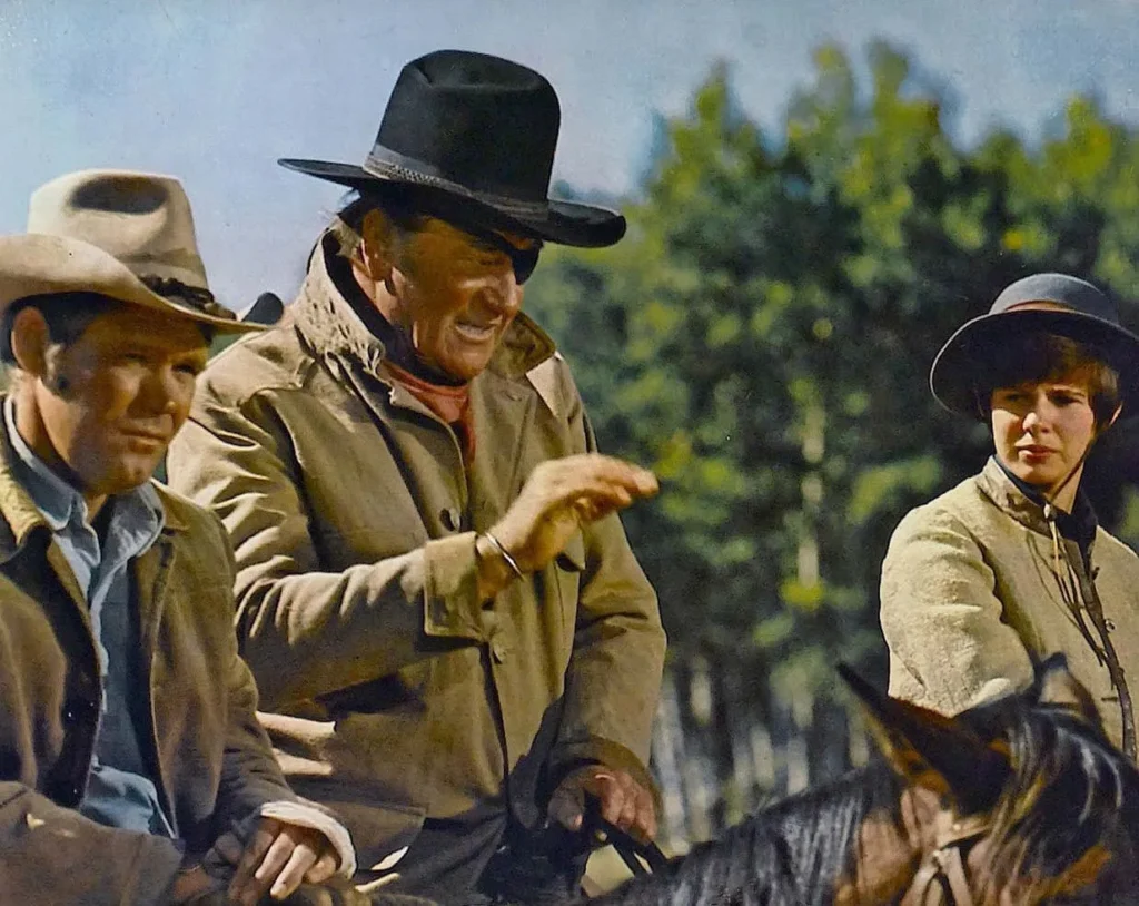 A group of three people from a scene in the classic Western movie “True Grit” (1969). They’re on horseback and dressed in period attire. The two men wear wide-brimmed hats, with the one in the center sporting a dark hat and a rugged jacket, laughing and pointing at something out of frame. The other man, to the left, is wearing a lighter hat and a denim jacket, looking on with a slight grin. To the right, a young woman with a resolute expression wears a bowler hat and a structured coat. Behind them, a backdrop of greenery suggests a rural outdoor setting.