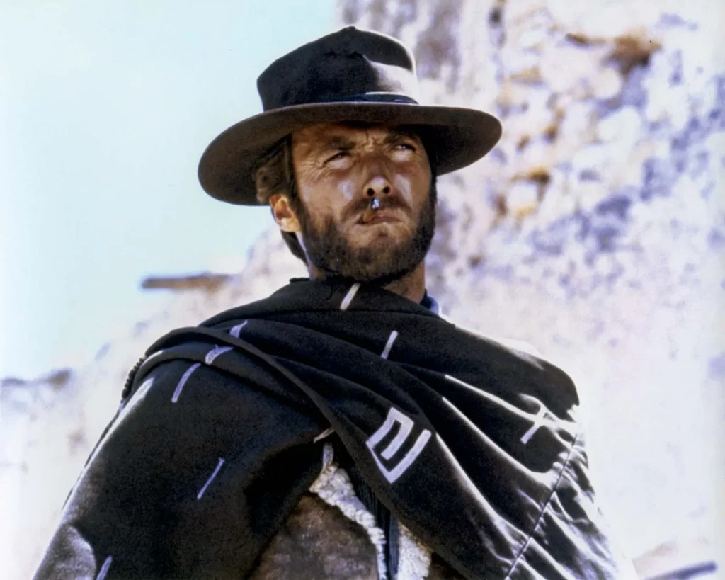 A rugged man with a stubbled face wears a dark cowboy hat and a poncho marked with white patterns over a traditional Western outfit. He has a stoic expression and is gazing off into the distance with an air of resolve. The man has a cigarillo clamped in his mouth, adding to his tough and ready-for-anything demeanor. The blurred background suggests a dusty, rocky terrain. This is a shot from the classic Western movie “A Fistful of Dollars” (1964).