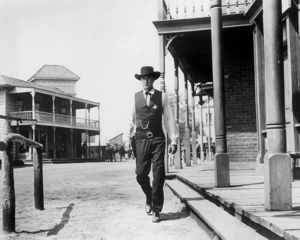 A lone lawman stands in a classic Western town, characterized by its wide dirt street and wooden buildings with a raised sidewalk. He's wearing a suit and vest with a sheriff's badge, a tie and a holster with a gun. His determined expression and the way he is walking towards the camera indicates that he is about to face a significant event from the movie “High Noon” (1952).