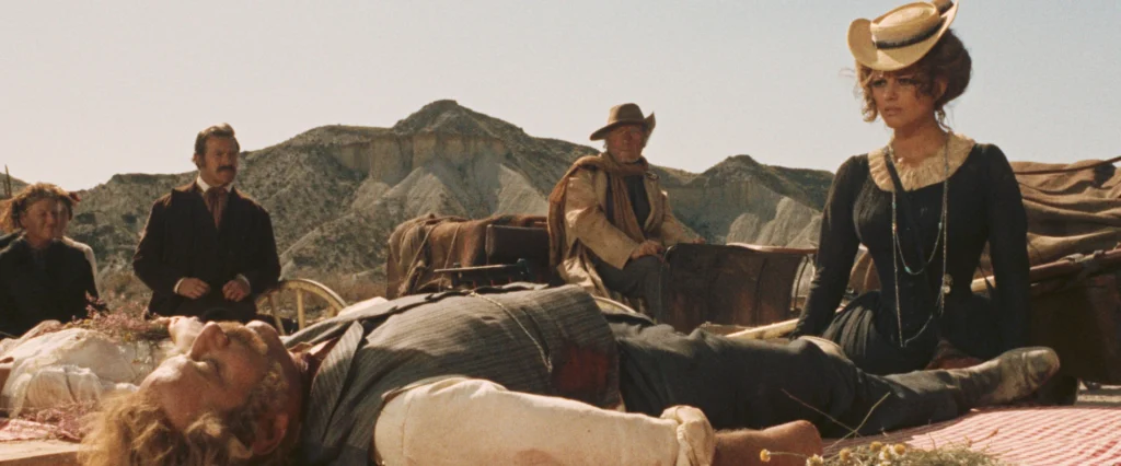 A dramatic scene from the movie “Once Upon a Time in the West” (1968) set against a backdrop of rugged hills. A man lies on a makeshift bed outdoors, appearing lifeless, which suggests he’s a casualty of a frontier conflict. Standing to his left is a stoic woman in mourning attire, and a well-dressed man with a mustache looks on solemnly. To the right, seated on the ground, is an elegantly dressed woman with a striking hat, her posture reflecting resignation or contemplation. A figure in a cowboy hat watches over the scene from behind a wagon. The setting is desolate and open, emphasizing the harshness of the environment and the narrative at play.