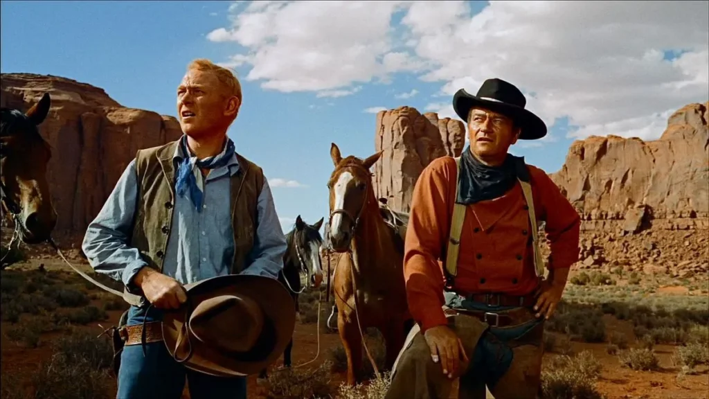 Two men in the vast expanse of the American Southwest, standing beside their horses. The man on the left, with a light blue shirt and a bandana, is holding his hat and looking off into the distance with an expression of concern or contemplation. The man on the right, wearing a dark hat and a rust-colored shirt, looks on earnestly, suggesting a pause during a challenging journey. The arid desert landscape with towering red rock formations hints at the epic scale of the movie this shot is taken from, “The Searchers” (1956).