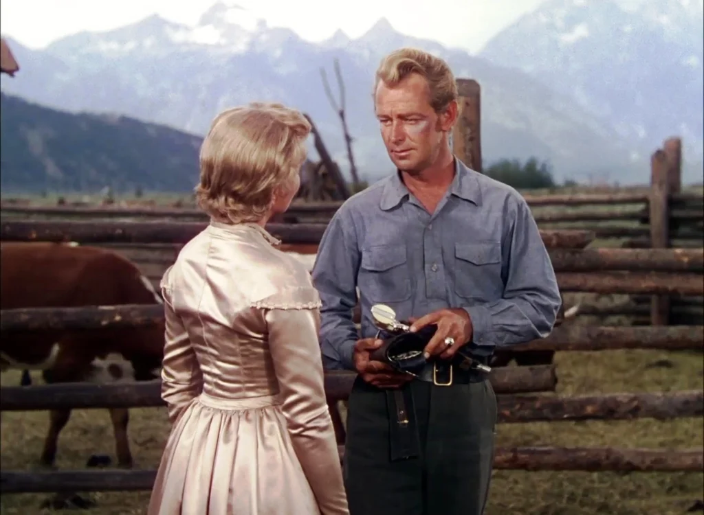 A poignant moment between a man and a woman in a pastoral setting from the Western movie “Shane” (1953). The man, dressed in a workman's blue shirt and holding a saddle, gazes thoughtfully at the woman, who is wearing a period-appropriate satin dress with lace trimmings, indicative of the 19th-century American frontier. They are engaged in a serious conversation, set against the backdrop of a rustic wooden fence and cattle grazing, with majestic mountains rising in the distance.