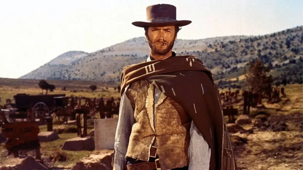 A rugged man stands in the foreground, epitomizing the quintessential Western gunslinger. He wears a brimmed hat and a poncho over a collared shirt, gazing intently beyond the frame. The background reveals a sparse, arid landscape typical of the American West, dotted with wooden grave markers and a wagon, suggesting a desolate cemetery. The stark, open terrain highlights the solitary figure, evoking the harshness and danger of frontier life. This is a shot from the movie “The Good, the Bad and the Ugly” (1966), and the character shown is the Man with No Name, played by Clint Eastwood.