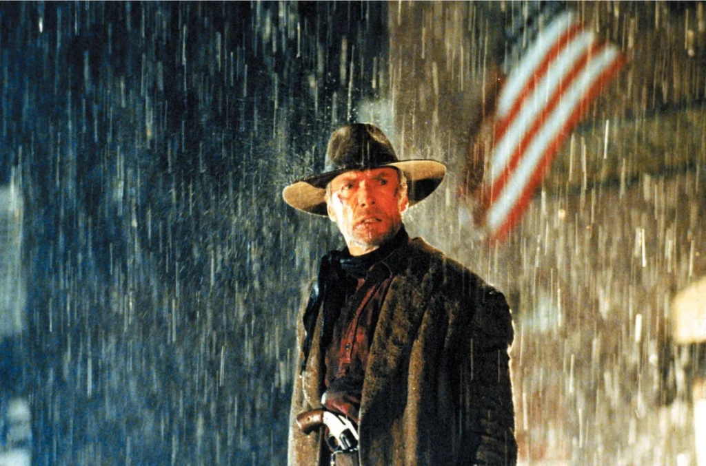 A rugged man in a rain-soaked Western setting, creating a somber and intense mood. He is wearing a cowboy hat and a heavy coat. His stern expression and focused gaze convey a sense of resolve or impending action. Behind him, the blurred American flag waving in the storm adds a layer of depth to the scene. The falling rain shrouds the setting in a veil of bleakness, emphasizing the gravity of this scene from the movie “Unforgiven” (1992).