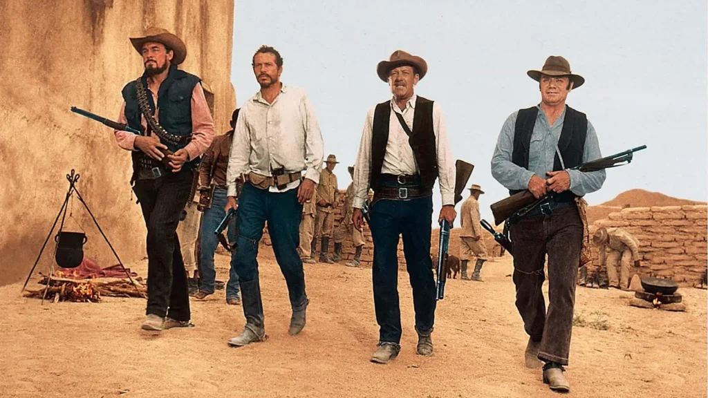 A shot from the movie “The Wild Bunch” (1969). It shows four men who exude the gritty essence of the Old West, each armed and dressed in the distinctive style of the era. From left to right, they carry rifles and wear hats, vests and boots, with the desert landscape and an adobe structure in the background underscoring the Western setting. A campfire with a boiling pot suggests a temporary stop in their travels. The stoic expressions and confident stances of these men convey a narrative of resilience and determination typical of classic Western films.