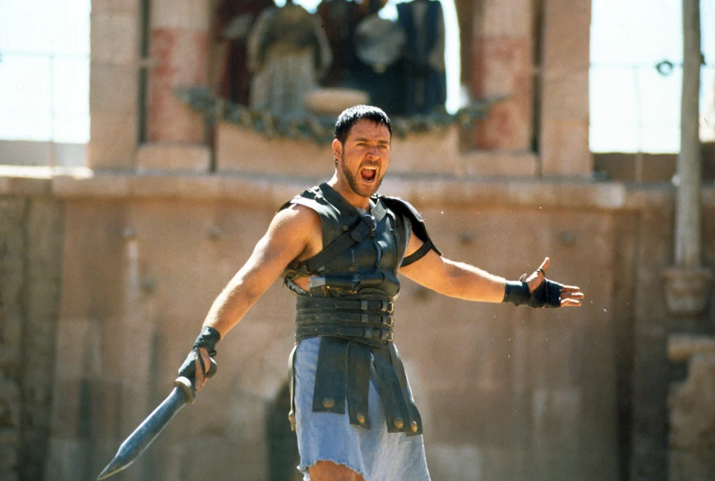 A man in ancient Roman gladiator attire, standing in an arena. He is wearing a leather armored sleeveless tunic, and his arms are outstretched in an impassioned gesture. He has a determined and defiant expression on his face, his mouth open as he’s speaking emphatically to the crowd. In one hand, he holds a short sword. This is an image from the movie “Gladiator” (2000), from the scene where Russell Crowe shouts his famous line: “Are you not entertained?!”