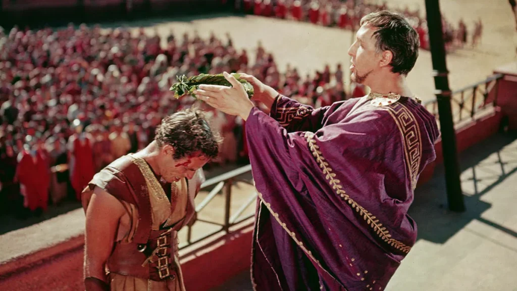 In the foreground, a man adorned with a laurel wreath is dressed in a purple toga with gold trim, signifying high rank. He extends his arm in a gesture that seems to bestow a laurel wreath upon the other man. This second man appears exhausted or injured, with a bloodied face, and is dressed as a gladiator. Behind them, a blurred crowd in red garments fills the stadium. This is a shot from the famous movie “Ben-Hur” (1959).