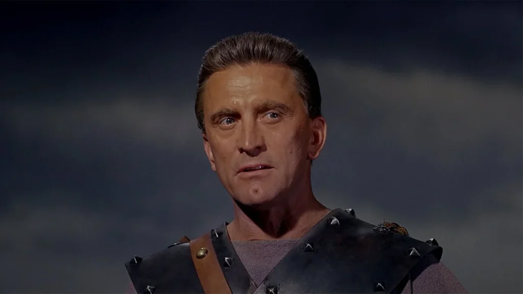 A close-up of a man in a contemplative or solemn moment. He is wearing leather shoulder armor with metal studs. His mature features, steady gaze and the set of his mouth convey a sense of experience and gravitas. The backdrop is a simple, cloudy sky. This is Kirk Douglas playing the title character of the movie “Spartacus” (1960).