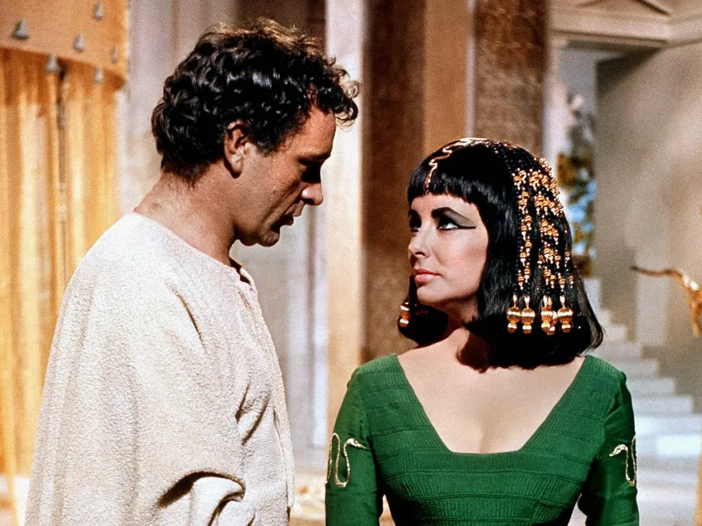 A man and a woman engaged in a close conversation. The man is dressed in a simple, off-white robe, his posture and facial expression imply earnestness or intensity in the dialogue. The woman wears an elaborate dark green dress with golden accents and a distinctive beaded headdress. Her makeup and hair reflect the style of ancient Egyptian nobility. The pair appear to be in a grand interior, with columns and luxurious decor in the background. This is a vibrant and colorful still from the movie “Cleopatra” (1963).