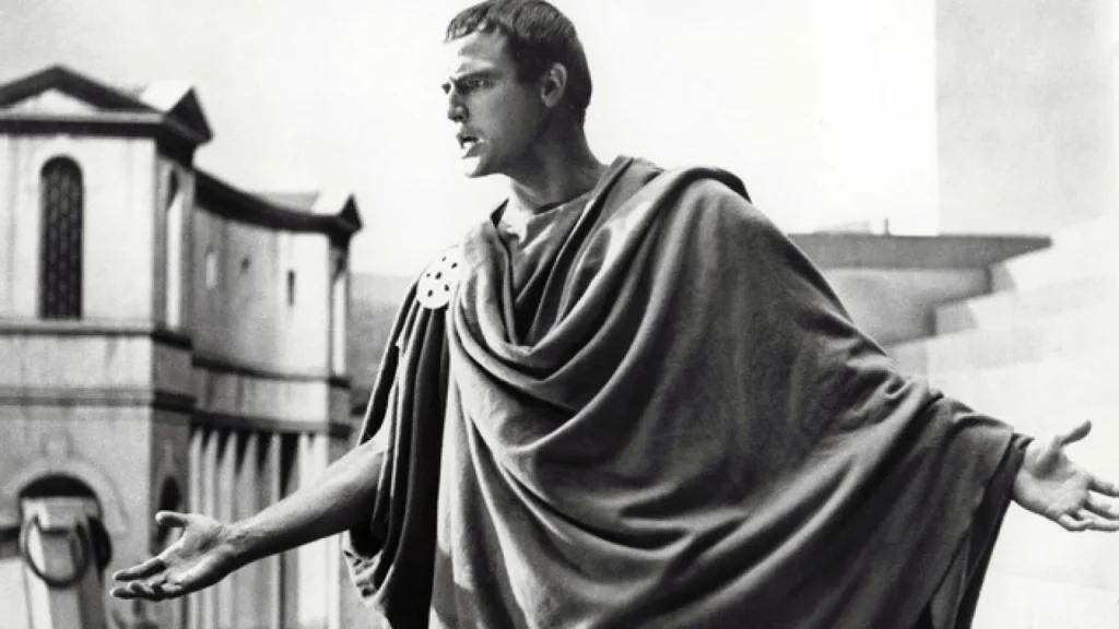 A man mid-speech or exclamation. He's dressed in a traditional Roman toga. His intense expression and the open, emphatic gesture of his hands convey a strong sense of emotion or persuasion, as if he is addressing a public assembly or engaging in a dramatic monologue. This is Marlon Brando playing Mark Antony in the movie “Julius Caesar” (1953).