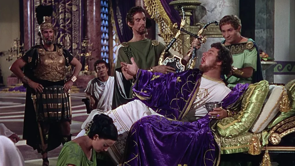 A vibrant image from the movie “Quo Vadis” (1951). The central figure, a man dressed in luxurious purple and gold, lounges on an ornate golden chaise with a lyre in his hand. His dramatic expression conveys a sense of indulgence. Surrounding him are several other characters: a stoic man in soldier's armor, individuals in togas and a woman in a green tunic who is turned away in deference to the central figure. The background features classic Roman columns and elaborate decorations.