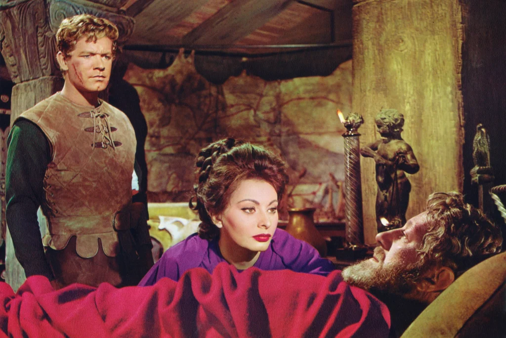 A young man in the left foreground stands with a solemn and attentive expression; he's clad in a leather tunic with a distinctive lacing at the collar. To his right, a woman with elaborate braided hair and wearing a rich purple dress leans over a figure lying on a bed, draped in a red blanket. She has a concerned and sorrowful expression as she looks upon the person in the bed, who has a graying beard and appears to be either ill or injured, judging by his reclined posture and the setting around him. The room is decorated with candlelight and sculptures. This is a shot from the movie “The Fall of the Roman Empire” (1964).