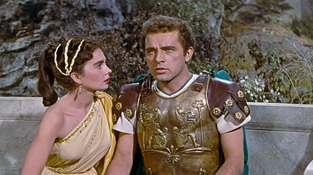 An intimate moment between a man and a woman dressed in luxurious ancient Roman attire. The woman, adorned with a golden headpiece and a flowing yellow dress, gazes at the man with a look of concern or questioning. The man, in detailed Roman soldier's armor embossed with regal designs, returns her gaze with a serious expression, hinting at a significant exchange or decision between the two. They are set against an outdoor backdrop within a palace garden or terrace. It’s a shot from the movie “The Robe” (1953).