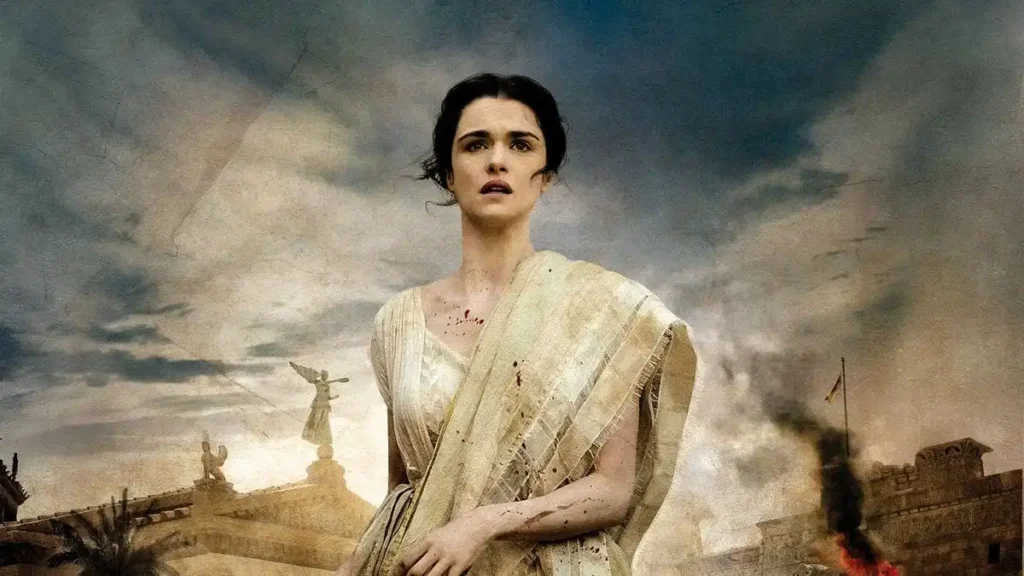 A woman dressed in ancient Roman-style clothing, standing against a backdrop of a dramatic sky filled with stormy clouds. Her attire, a white stola with delicate fabric, suggests she may be of high status or nobility. The woman has a look of resolve or concern on her face, which contrasts with the smudges of dirt and bloodstains on her dress, indicating she may have been through a traumatic event. In the background, classical Roman architecture is visible, including statues and stone buildings, with a hint of smoke or fire near one structure, adding to the atmosphere of turmoil or disaster. This is a promotional image for the movie “Agora” (2009).