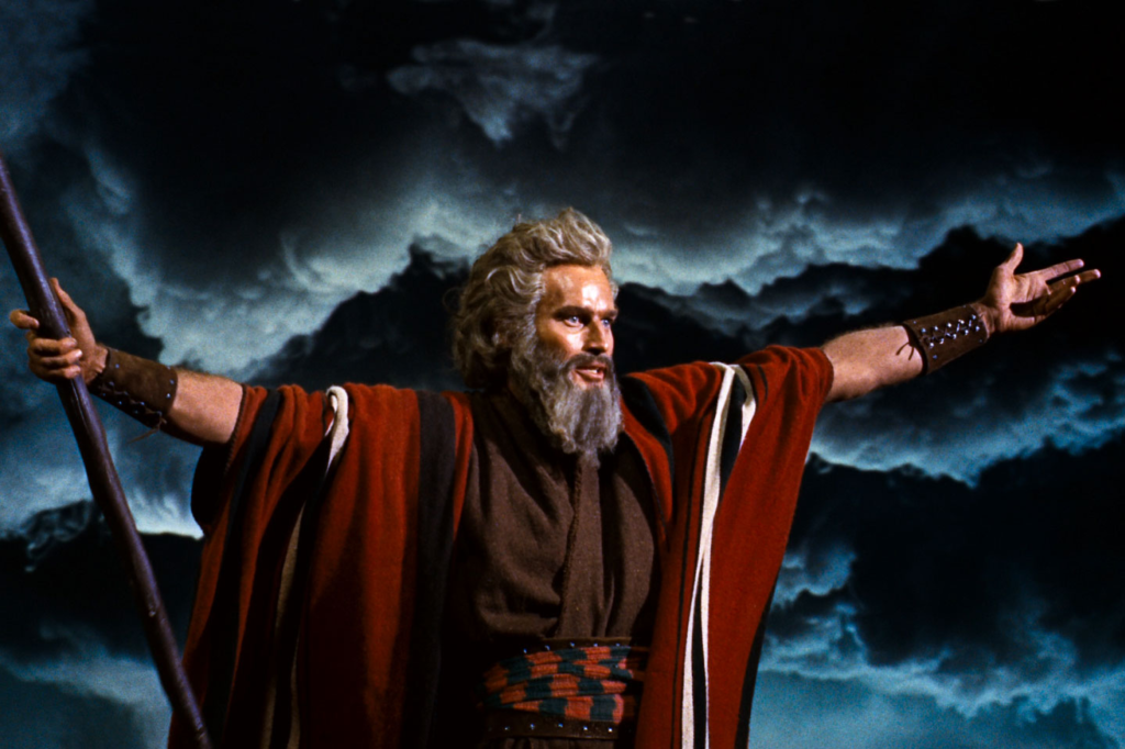 A moment of grandeur from the classic film “The Ten Commandments” (1956), featuring an iconic character portrayed as a wise and powerful leader. It’s Moses, and he stands with arms outstretched, a staff in one hand, against a dramatic backdrop of tumultuous clouds and dark mountains. His expression is one of command and determination, his face framed by a flowing beard and hair, suggesting his sage-like wisdom and authority. The character's costume, a vibrant red cloak over a more subdued tunic, emphasizes his central role and the epic nature of the scene.