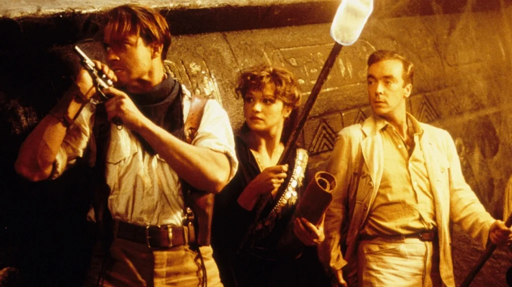 A scene from the action adventure movie “The Mummy” (1999). It features three characters in an ancient, dimly-lit tomb. The man on the left is poised for danger, gripping a pistol and looking intently ahead, his attire and equipment suggesting he's the rugged, adventurous type. The woman in the center, also alert and holding a torch, looks on warily, her attire a mix of practicality and period-specific elements, hinting at her role as both a scholar and an active participant in the adventure. The man on the right, dressed more formally, appears thoughtful and possibly skeptical.