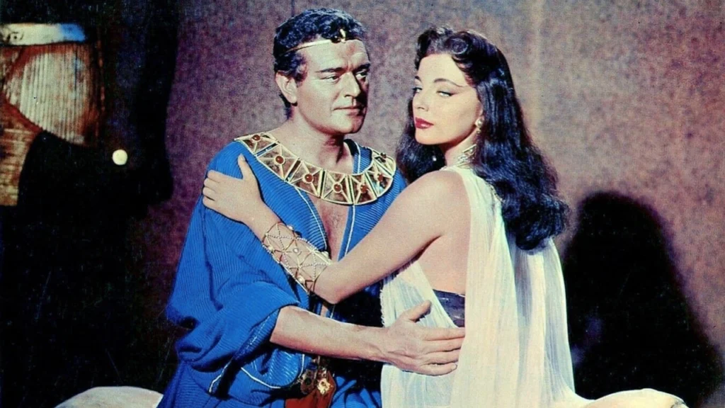 A romantic still from the classic film “Land of the Pharaohs” (1955). It depicts an intimate moment between two characters dressed in ancient Egyptian costumes. The man, adorned in a blue robe and golden jewelry that accentuates his royal status, gently embraces a woman who looks back at him with an expression of soft affection. She wears a flowing white gown and is embellished with delicate jewelry, enhancing her elegant and graceful appearance. The dimly lit background suggests a private, possibly secretive moment, adding to the dramatic and emotional depth of the scene.