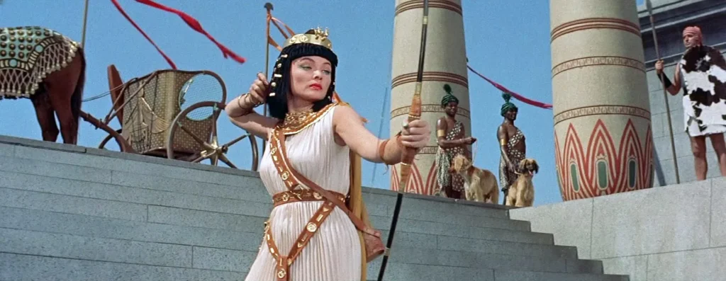 A vibrant scene from the classic film “The Egyptian” (1954). It showcases a striking image of a queen in Ancient Egypt, standing majestically on a grand staircase outside a temple. She is dressed in a traditional white gown accented with gold belts and ornaments and a royal headdress, and she holds a long, golden bow. The setting is opulent, with towering columns and vibrant flags, while attendants and animals, including horses and dogs, add to the ceremonial ambiance.