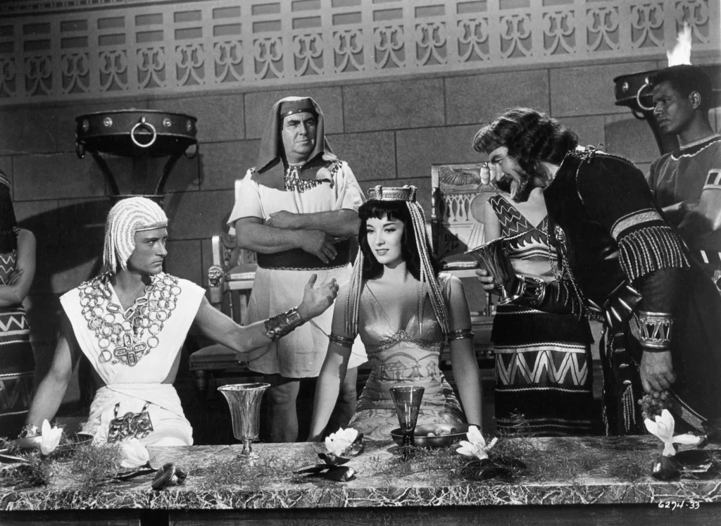 A scene from “The Pharaoh’s Woman” (1960), a classic black-and-white film set in Ancient Egypt. It features a group of characters in elaborate costumes, showcasing an opulent royal setting. On the left, a young man with a distinctive white headdress and a broad collar made of beads is seated at a table, turning towards a beautiful woman dressed as an Egyptian queen, complete with a ceremonial headpiece and intricate jewelry. She appears engaged and poised as she listens to him. Standing beside her is a man in a dark, ornate military uniform, holding a goblet and leaning in to join the conversation. On either end of the table, two more men in traditional Egyptian attire observe the exchange, adding to the scene’s dynamic. The background is adorned with Egyptian motifs.